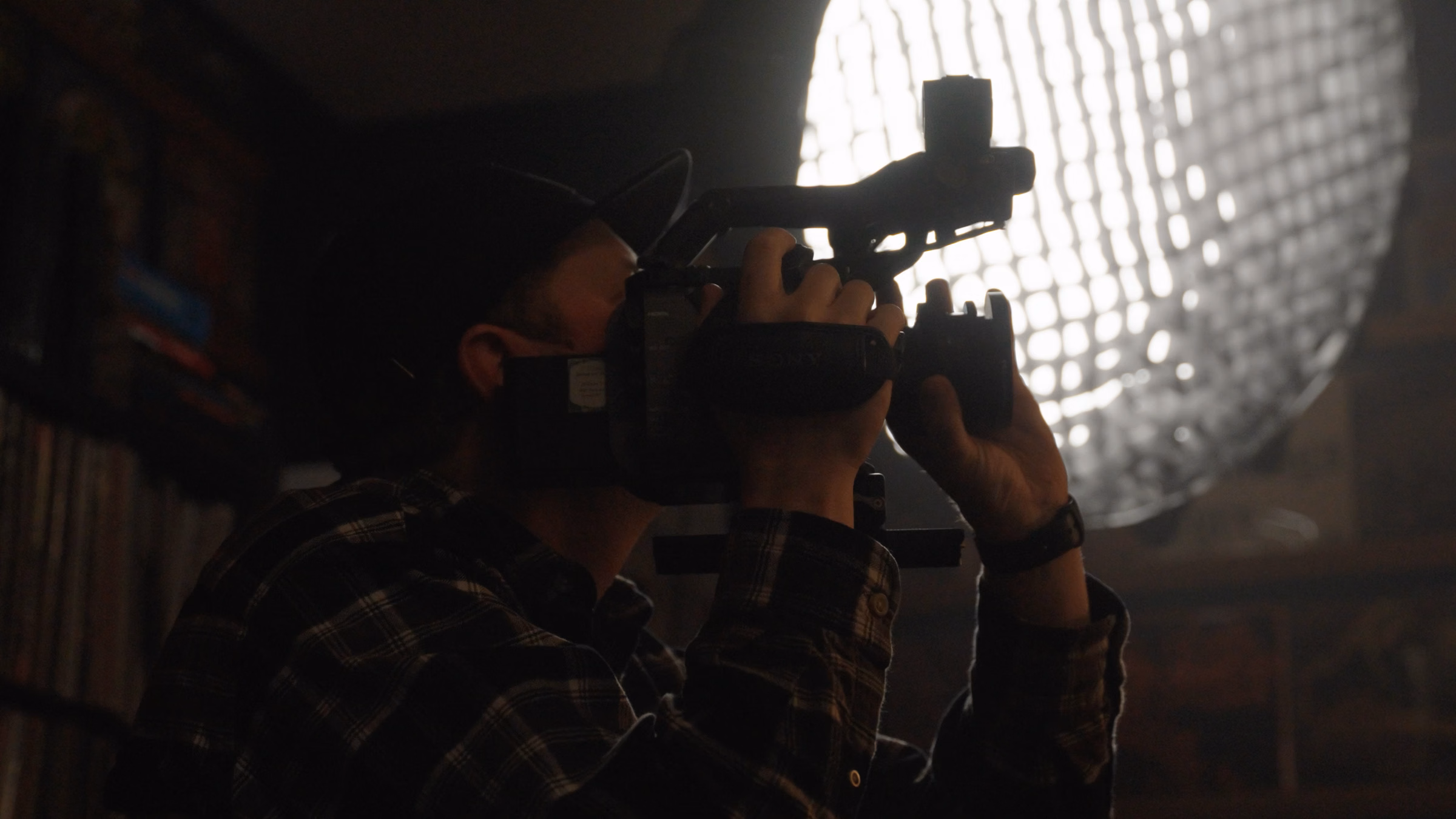 Person wearing a cap filming with a video camera in a dim room with bright light source behind.