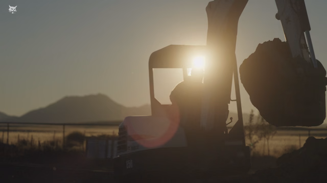 Silhouette of excavator digging at sunset with mountains in the background.