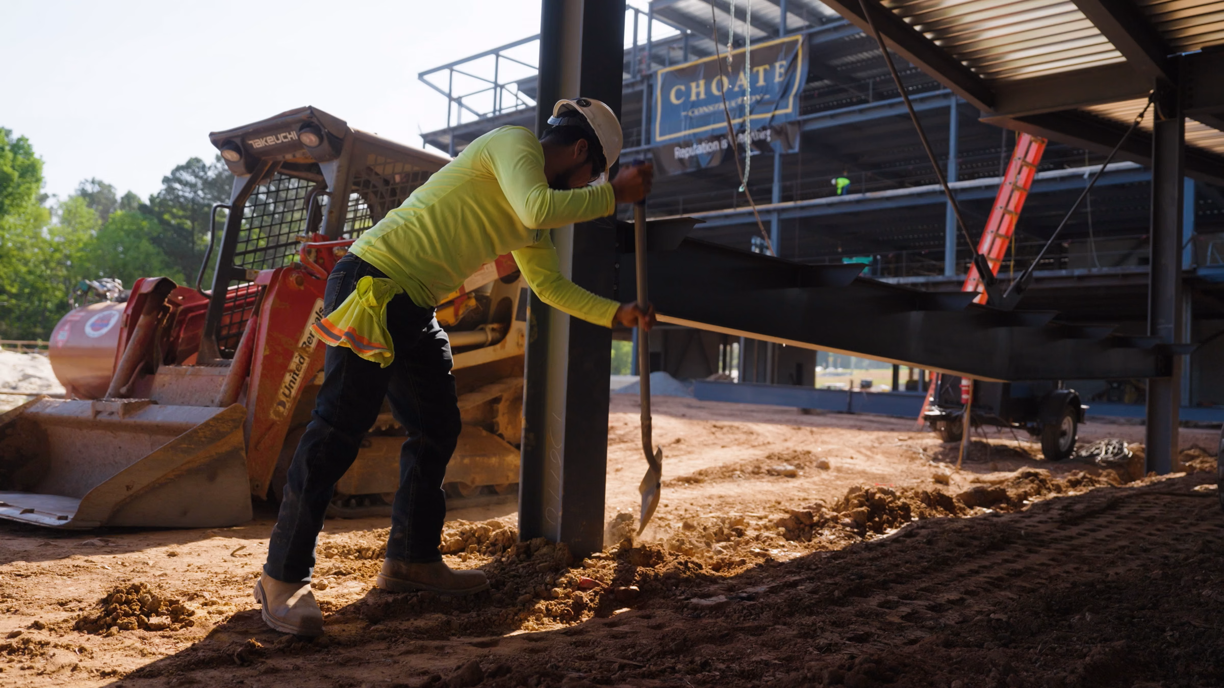Construction worker in hard hat shoveling dirt inside a building frame near heavy machinery.