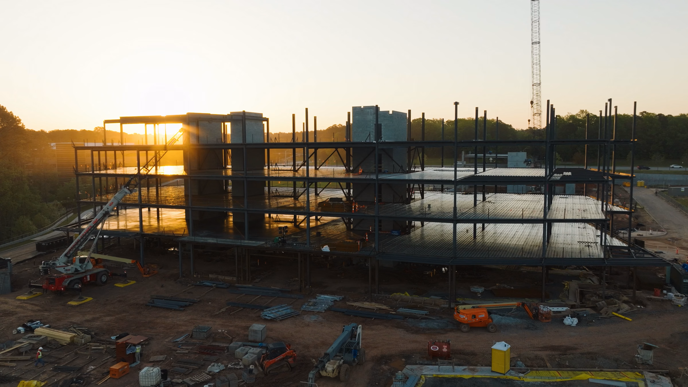 Steel framework of a multi-story building under construction at sunrise with heavy machinery around.
