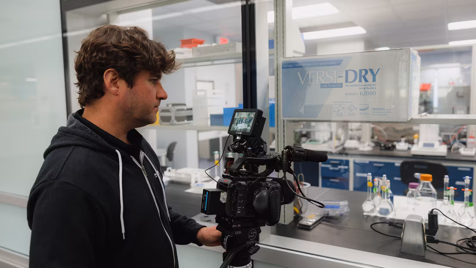 Man operating a professional video camera filming through a glass window into a lab with scientific equipment.