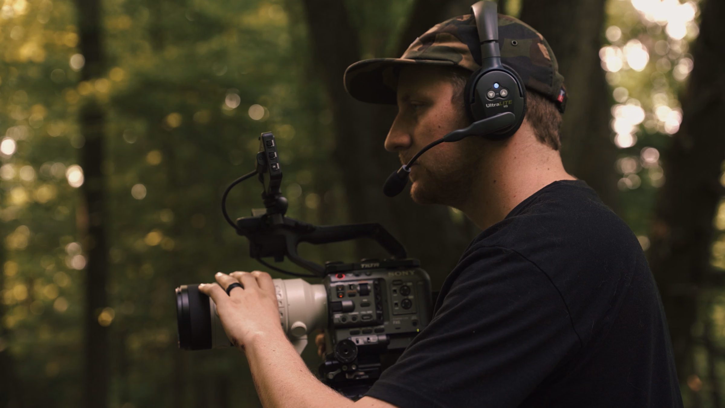 Man wearing headset and cap filming with professional video camera in outdoor wooded area.