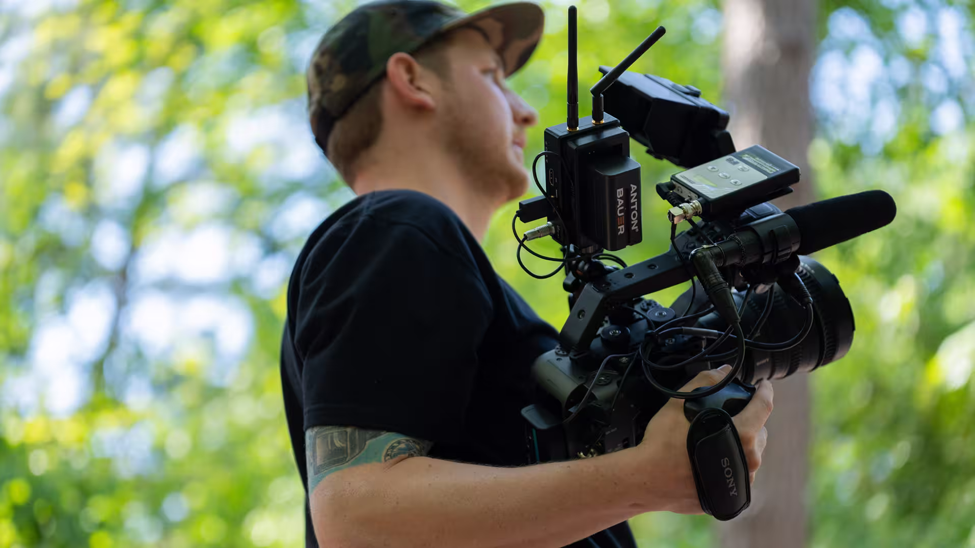Man in camo hat holding a professional video camera with audio equipment outdoors.