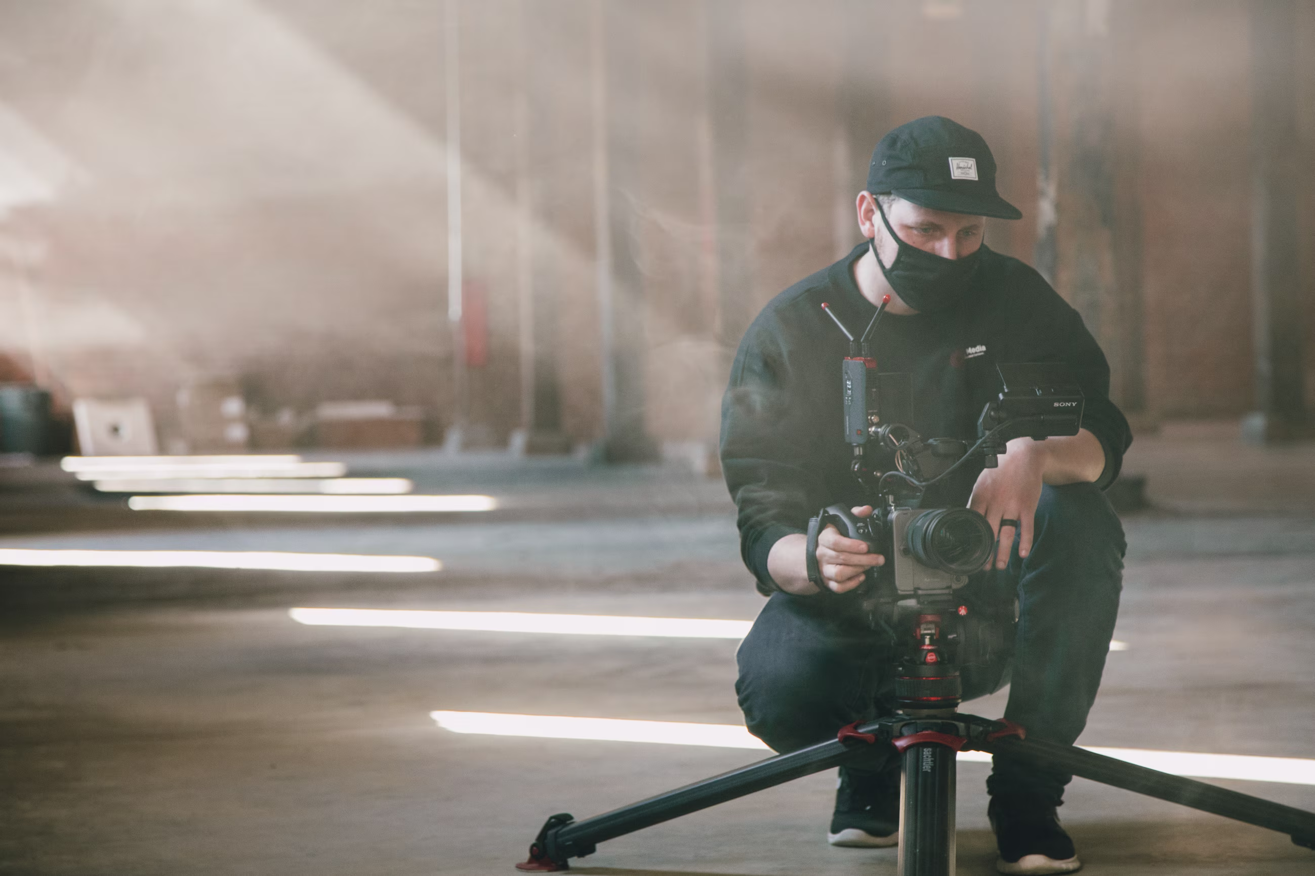 Man wearing black mask and cap operating professional camera on tripod indoors with light beams.