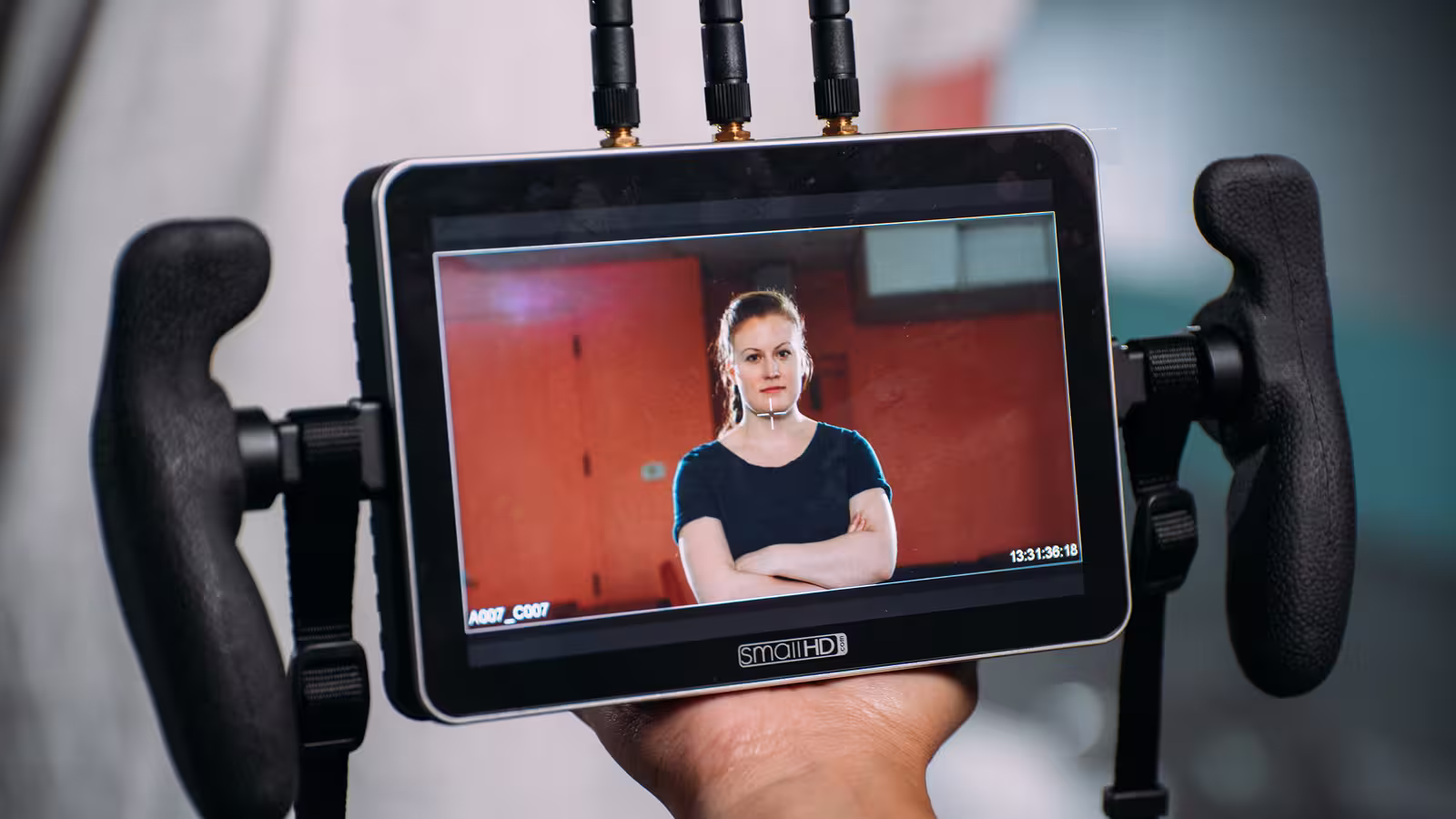 Hand holding a SmallHD monitor showing a woman in a black shirt with arms crossed on screen.
