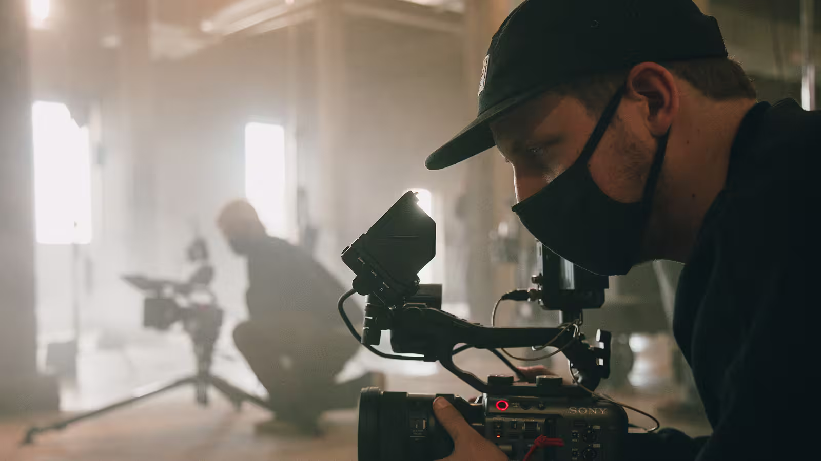 Filmmaker wearing a black cap and mask operating a Sony video camera indoors with a blurred figure in background.
