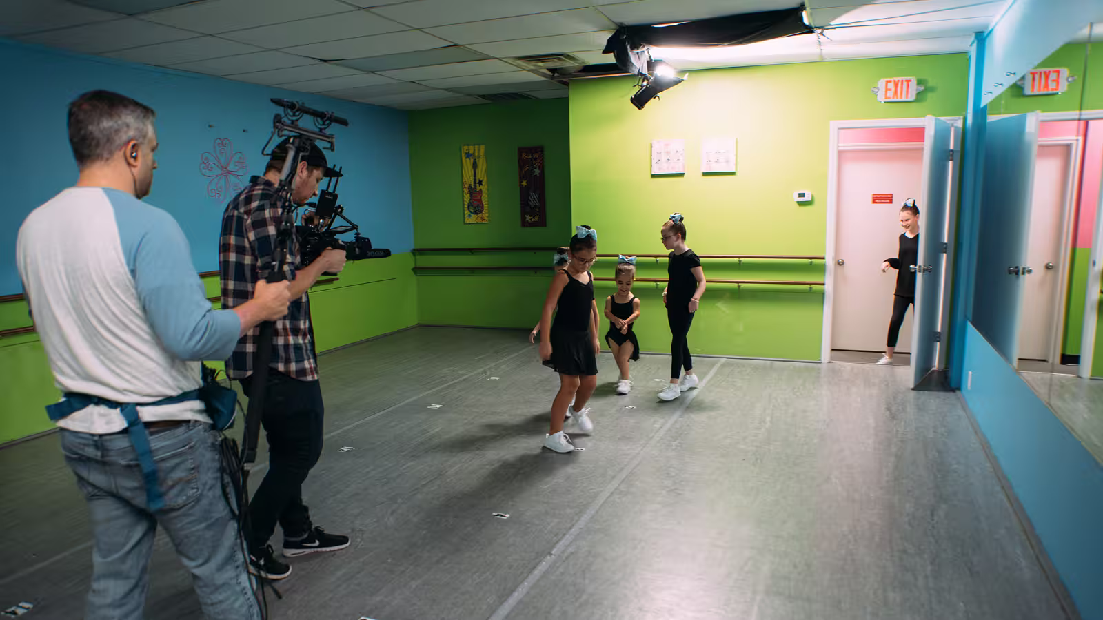 Three young dancers in black outfits practicing in a studio as two men film them.