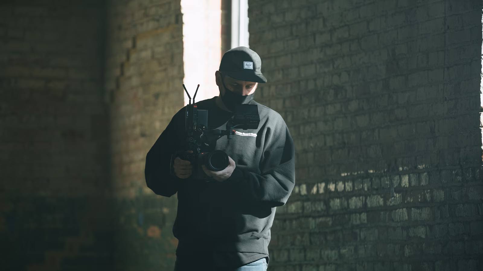 Man wearing cap and mask filming with a professional camera in a dimly lit brick-walled room.