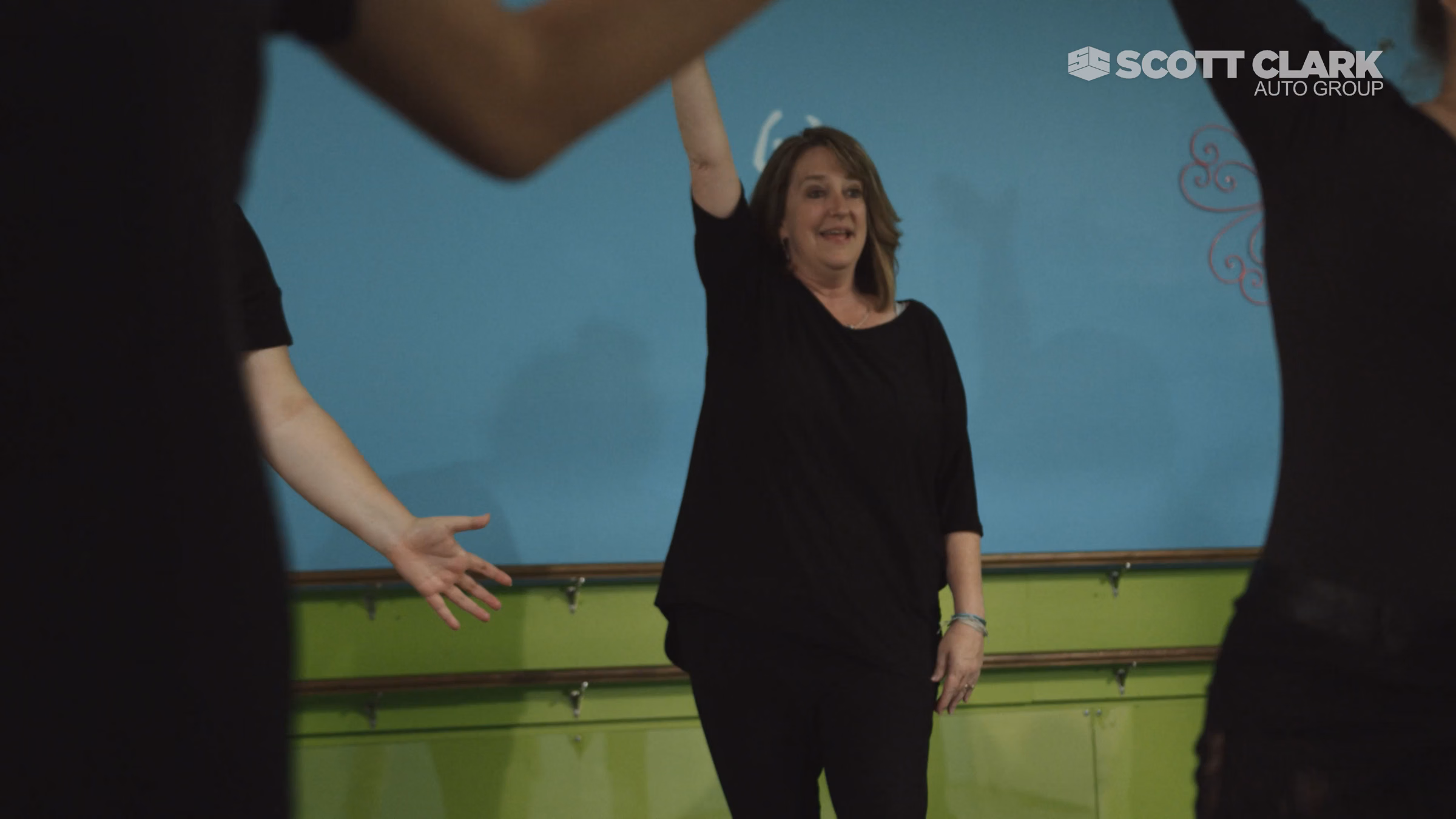 Smiling woman in black raising hand in a dance or exercise studio with others partially visible.