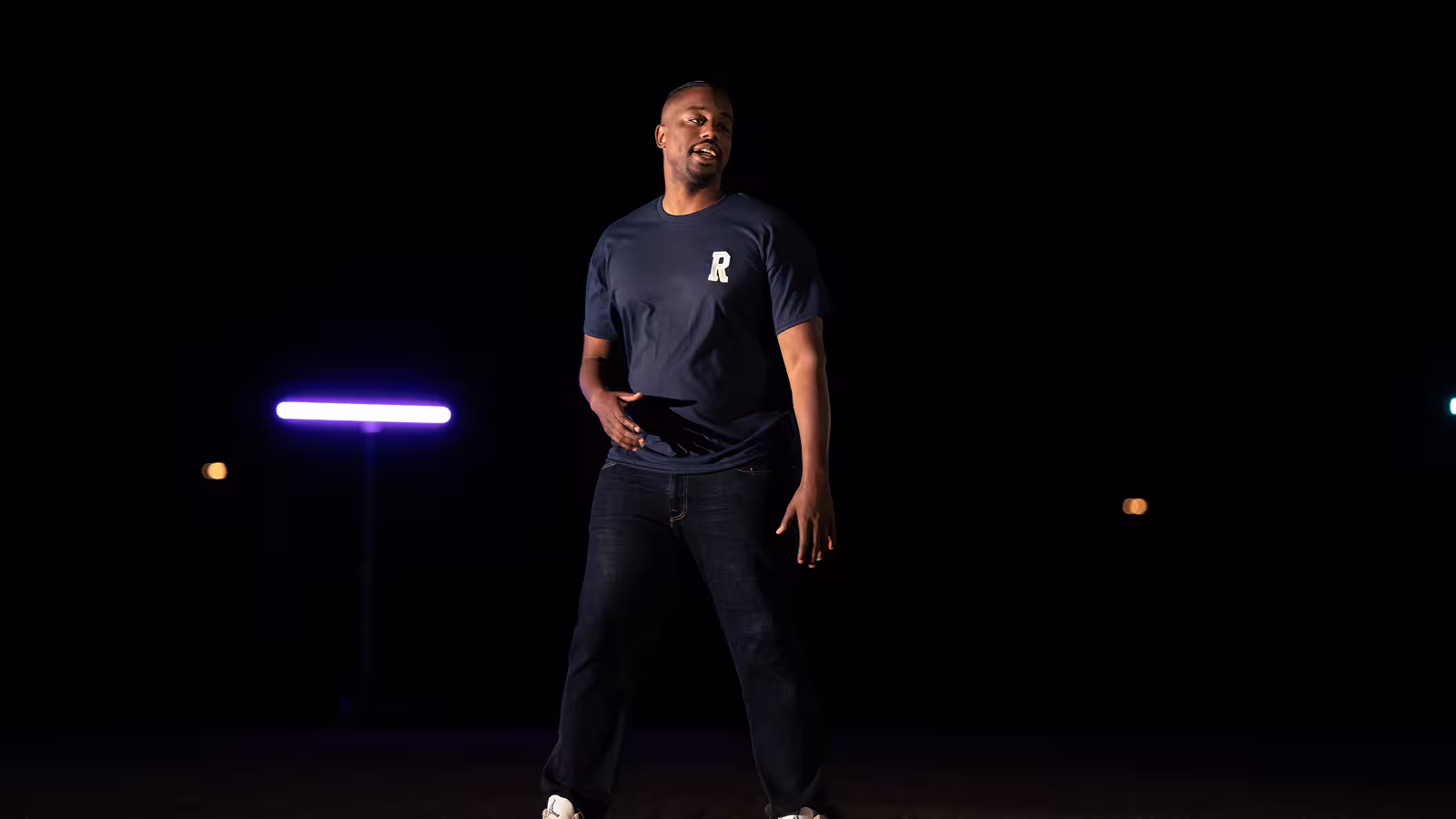Man in dark jeans and blue shirt with 'R' logo standing against dark background with purple light.