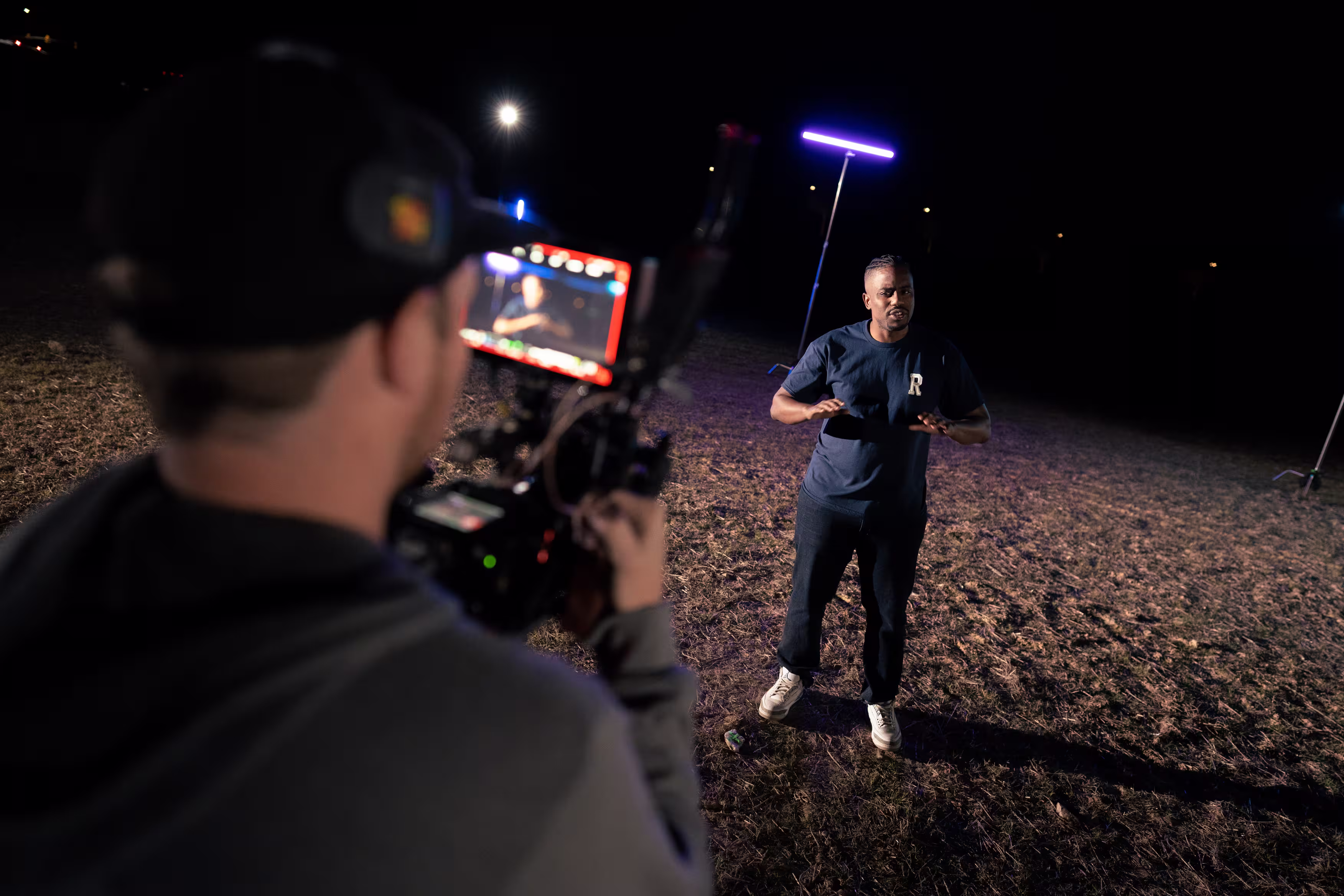 Man in dark shirt speaking in front of a camera operator outdoors at night with purple lighting.