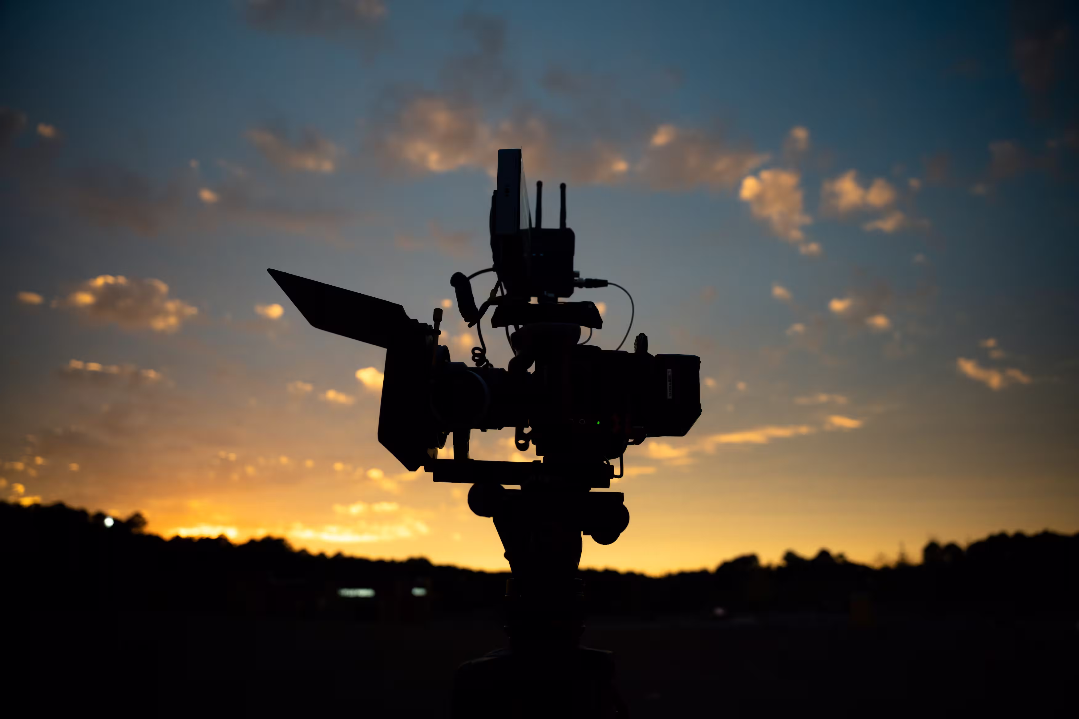Silhouette of a professional video camera against a sunset sky with scattered clouds.