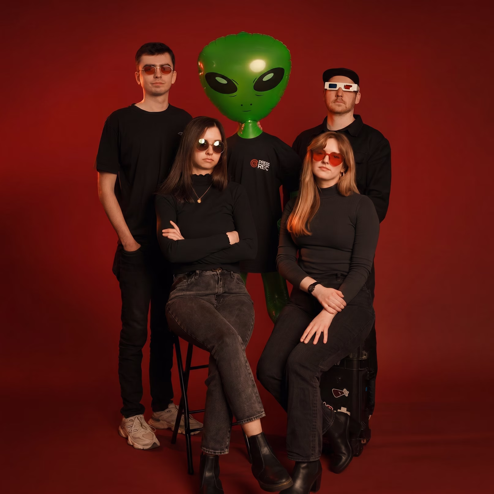 Five people wearing sunglasses pose against a red background with a large green inflatable alien.