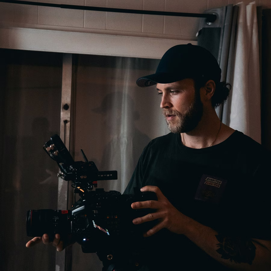 Man with black cap and beard operating a professional video camera indoors near a window with curtains.