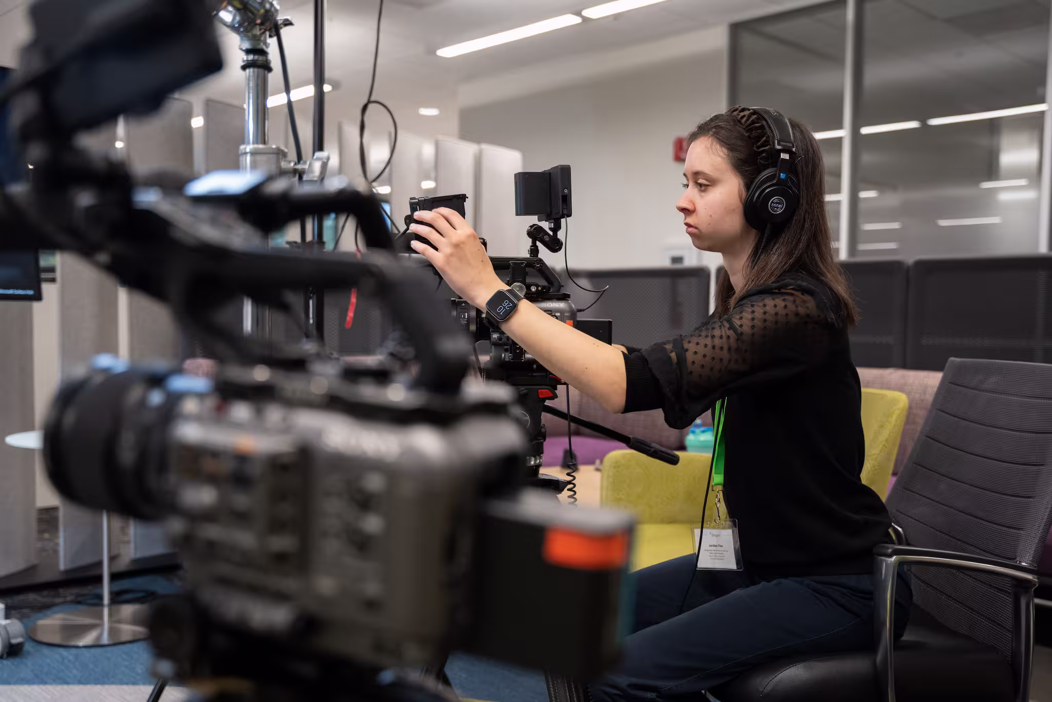 Woman wearing headphones adjusts professional video camera in an office setting.