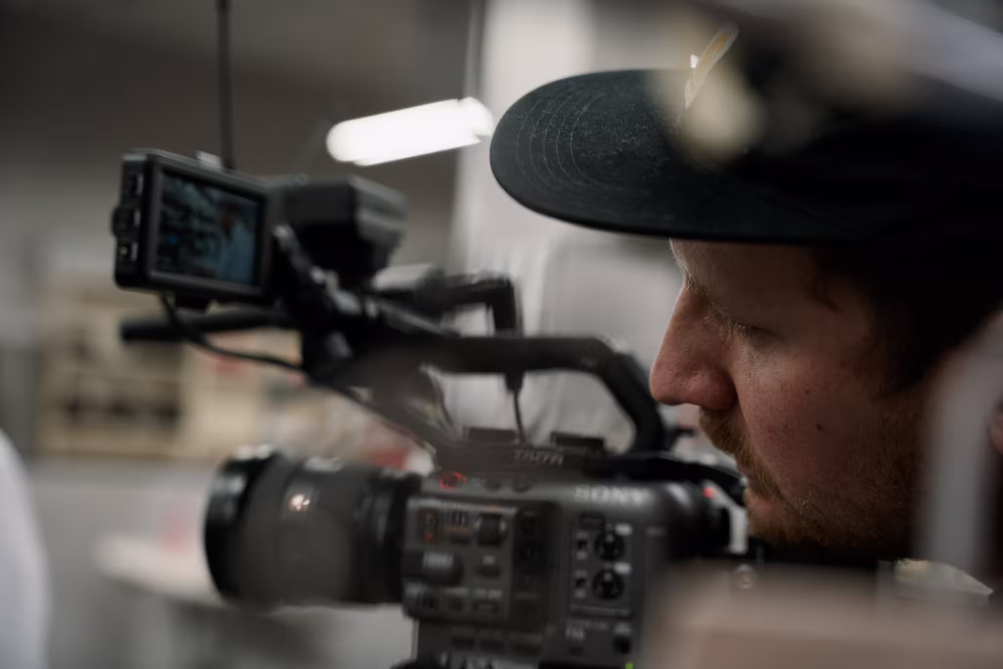 Man wearing black cap operating a professional video camera with attached monitor indoors.