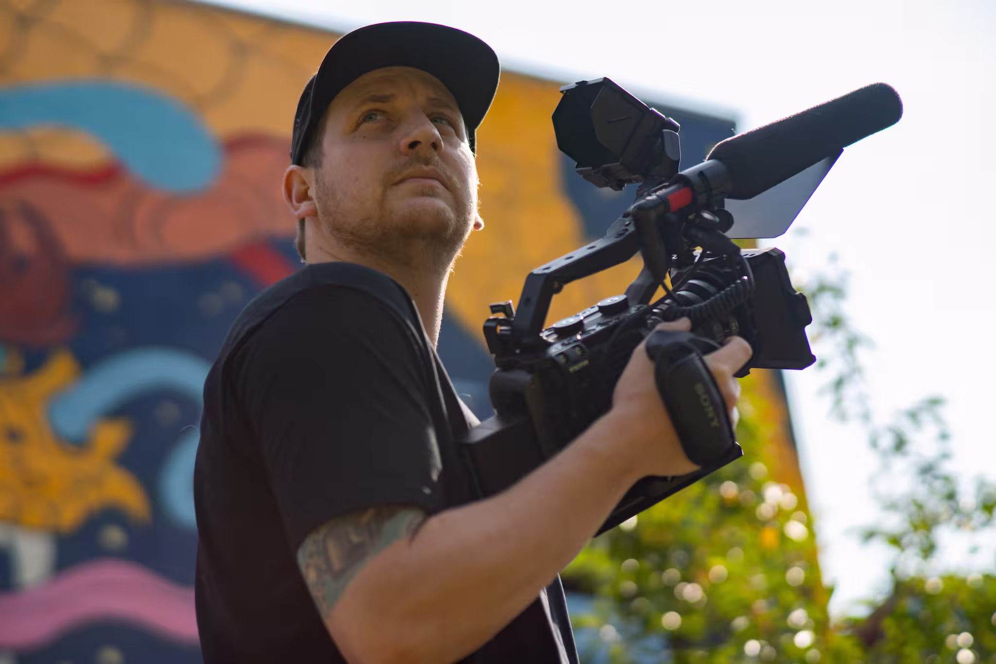 Man in black holding professional video camera with microphone outdoors near colorful wall art.