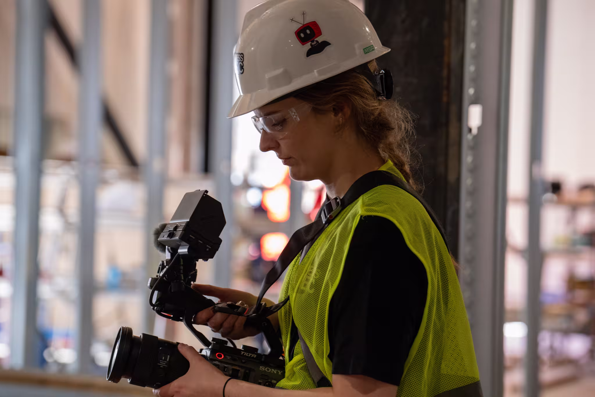 Person wearing a white hard hat and yellow safety vest operating a professional camera indoors.