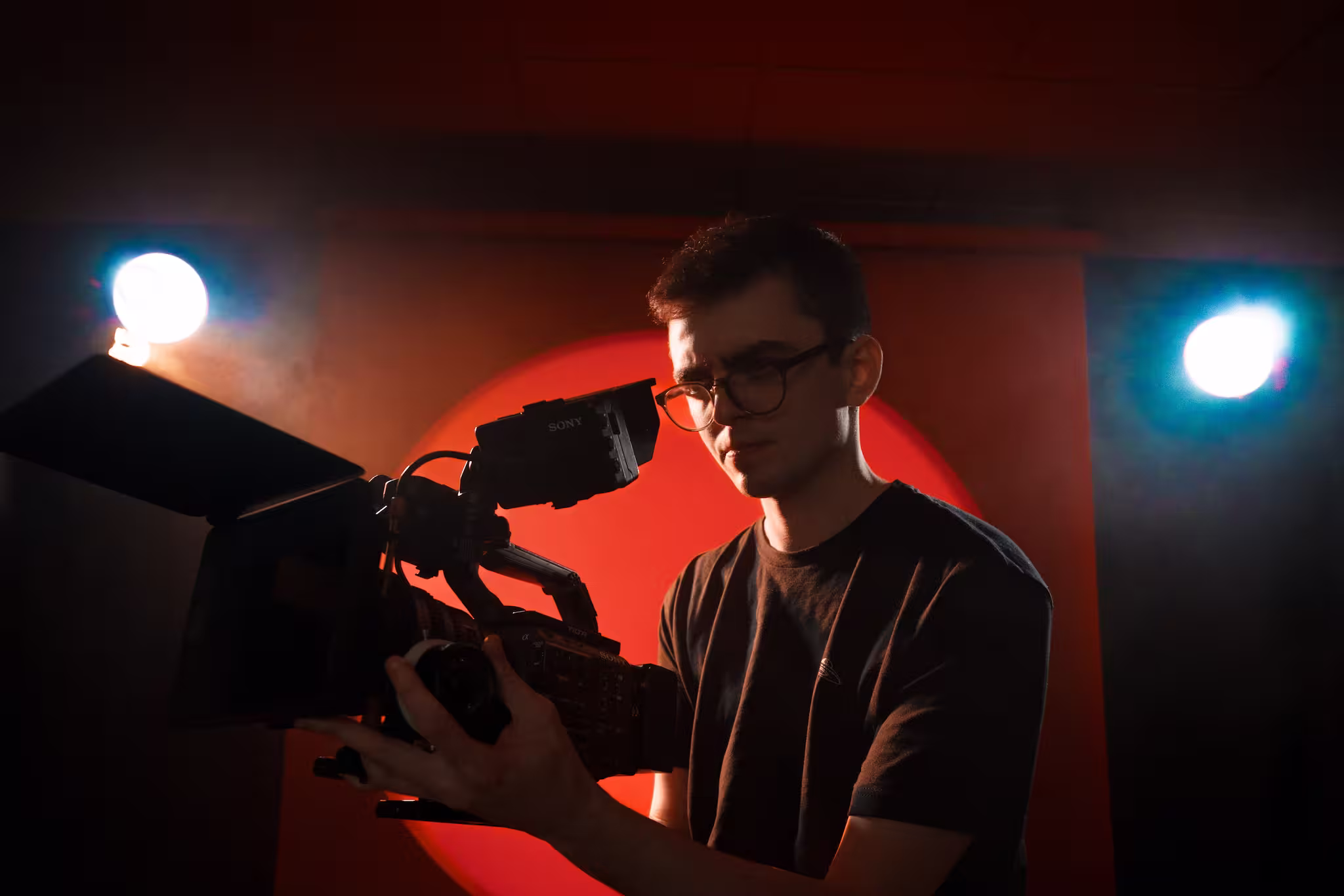 Man wearing glasses filming with a Sony video camera against red and black background with bright lights.