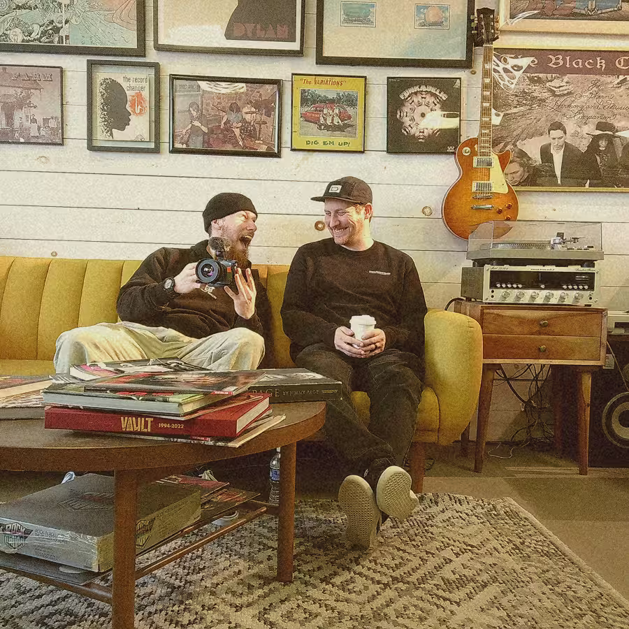 Two men sitting on a yellow sofa, one holding a camera, the other a cup, laughing with framed art and guitar behind.