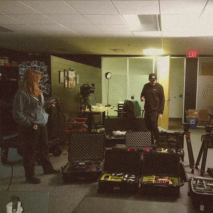 Two people in a dimly lit room with camera equipment cases and tripods on the floor.