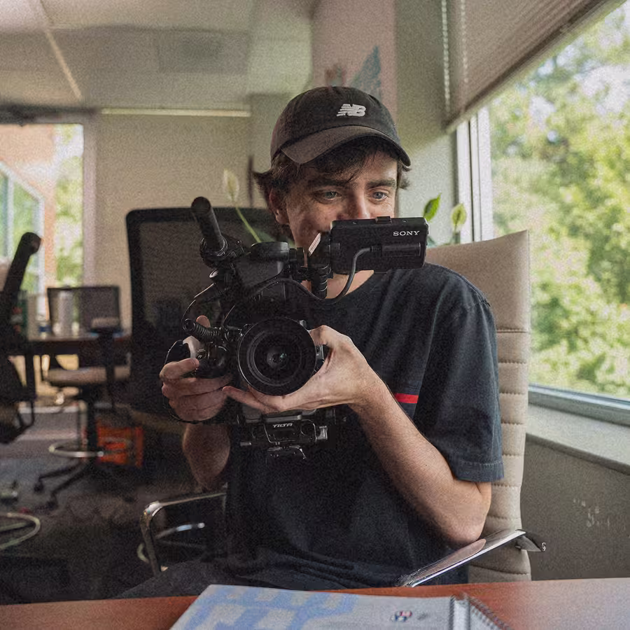 Man wearing a black cap holding a professional Sony video camera indoors near a window.