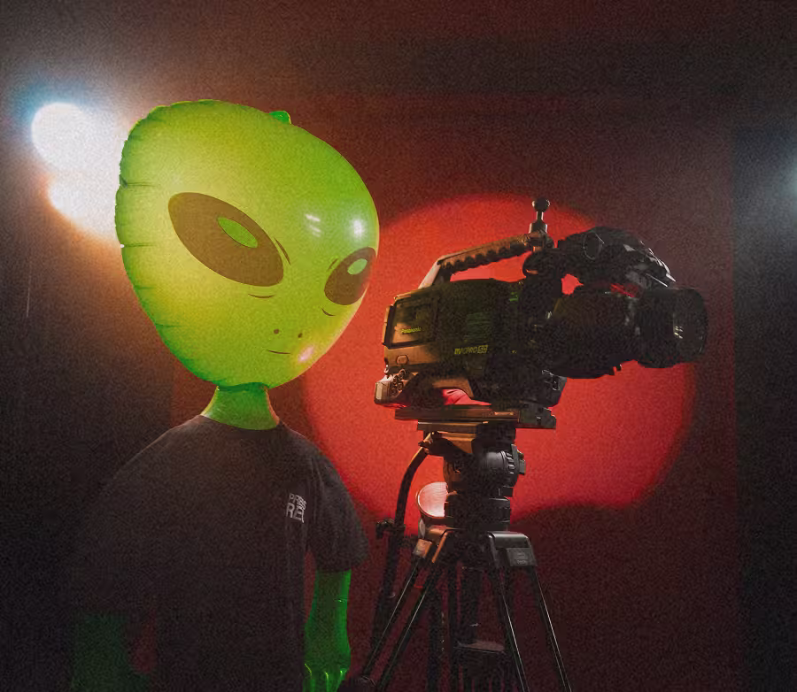 Person wearing large green alien mask stands beside professional video camera on tripod with red and white lights.