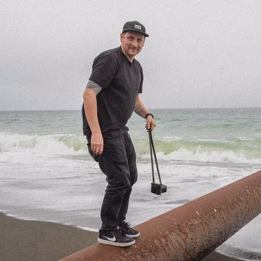 Man in black clothes balancing on a large rusty pipe at the beach while holding a camera.