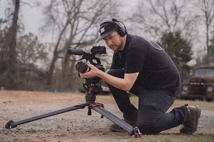 Man crouching outdoors filming with a camera on a tripod, wearing headphones and a cap.