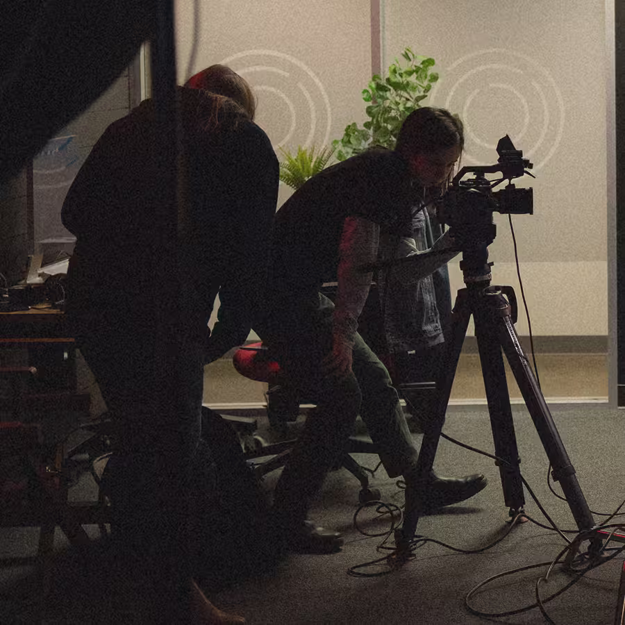 Two people setting up video camera on tripod in a dimly lit room with plants in background.