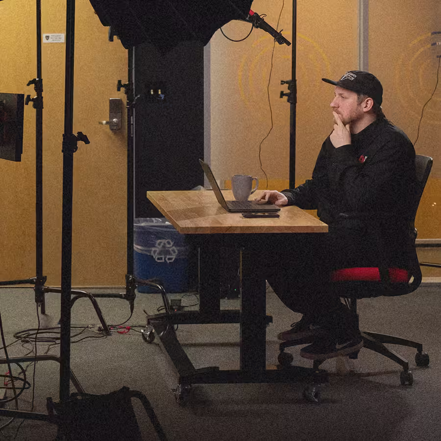 Man in black cap and clothes working on a laptop at a wooden table in a studio setting.