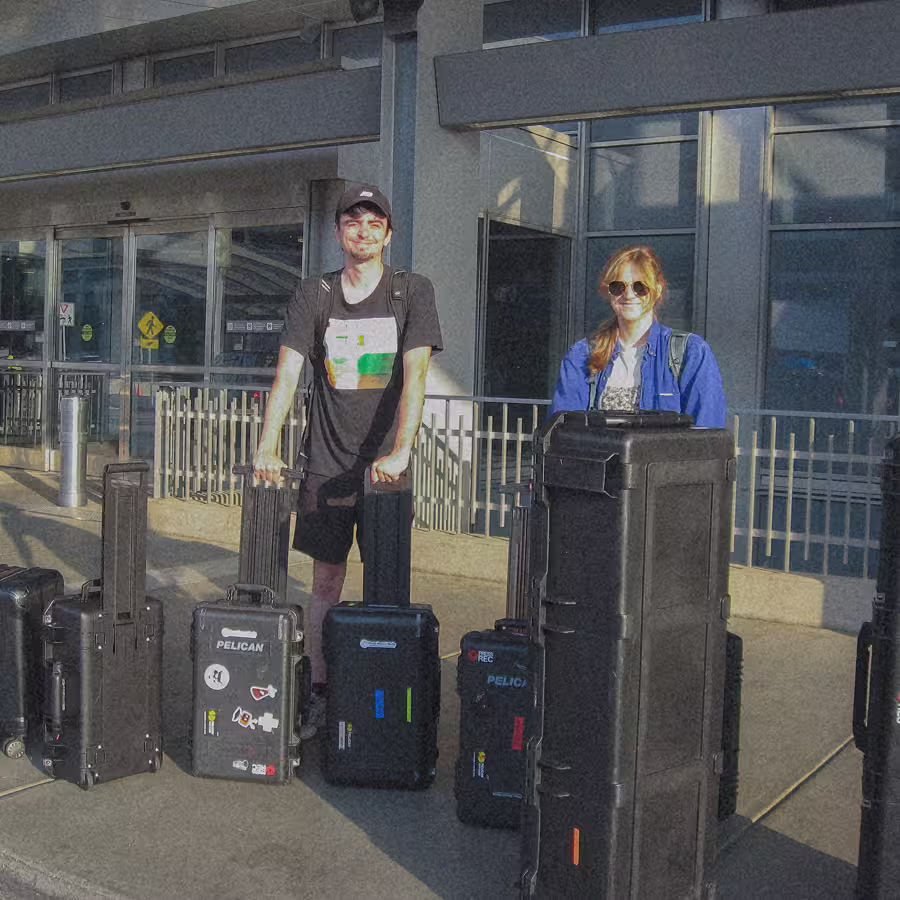 Two young adults with multiple large black hard-shell luggage cases outside a building entrance.