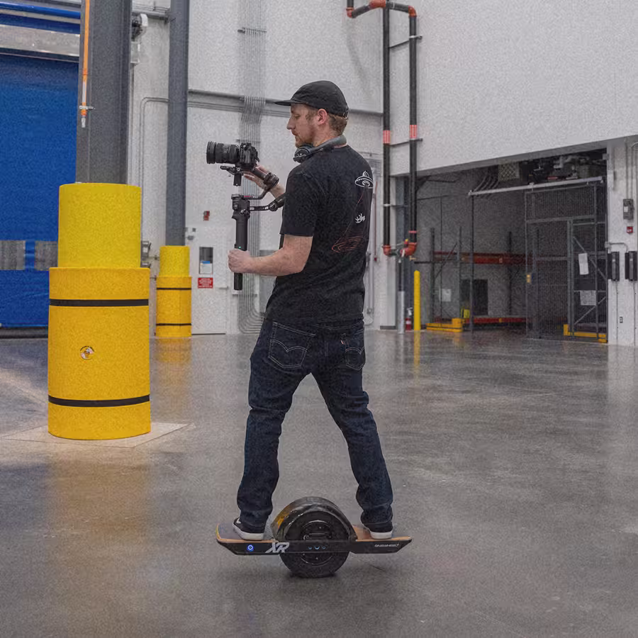 Man wearing black cap and t-shirt riding a one-wheeled electric board while filming with a camera rig.