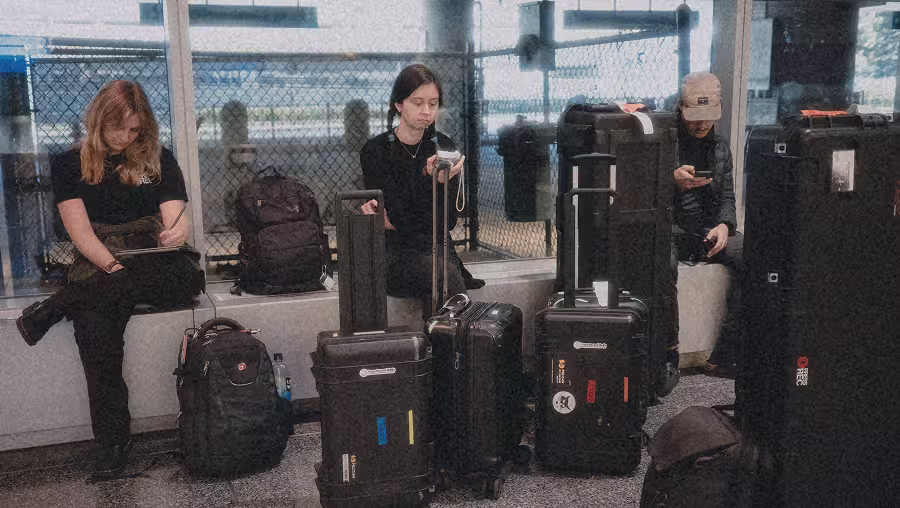 Three people sitting on a bench at an airport with multiple black suitcases and backpacks.