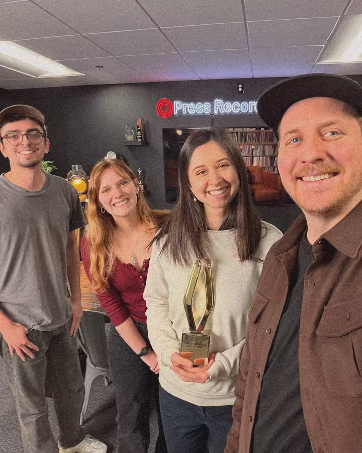 Four smiling people indoors, one holding a Viddy Award, with a 'Press Record' neon sign in background.