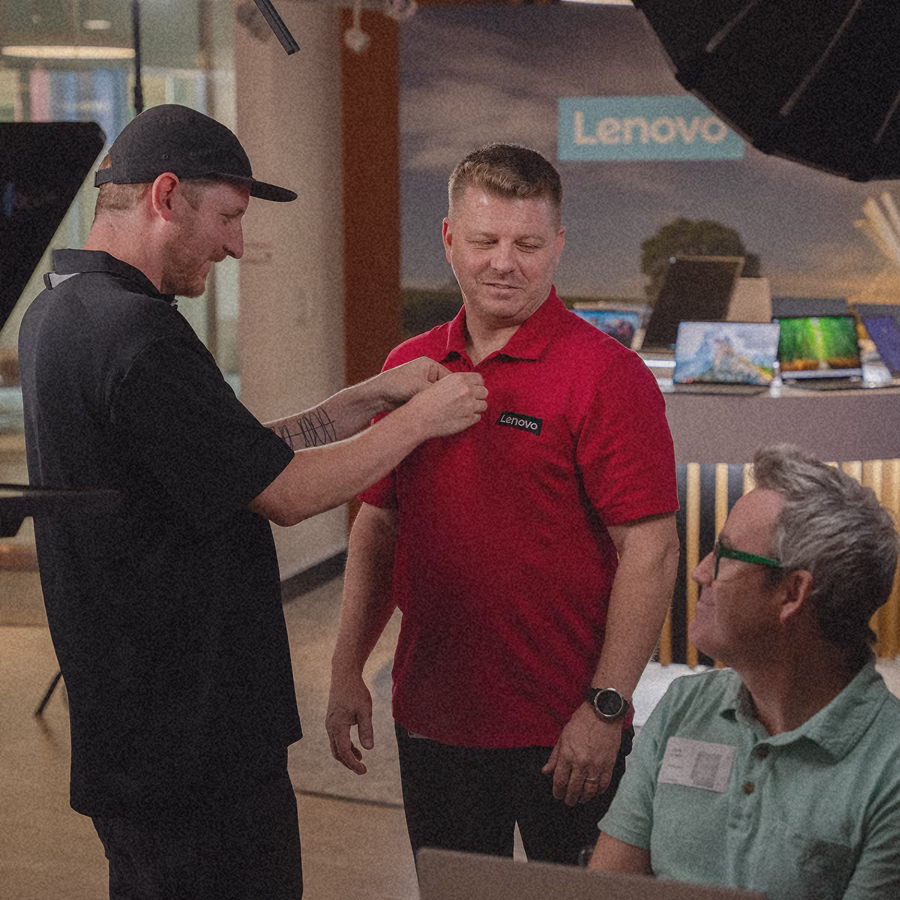 Man in black shirt attaching a microphone to a man in a red Lenovo shirt during a filming setup.