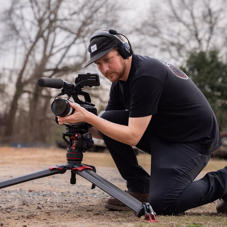 Man kneeling outdoors adjusting a camera on a tripod with a headset and cap on.