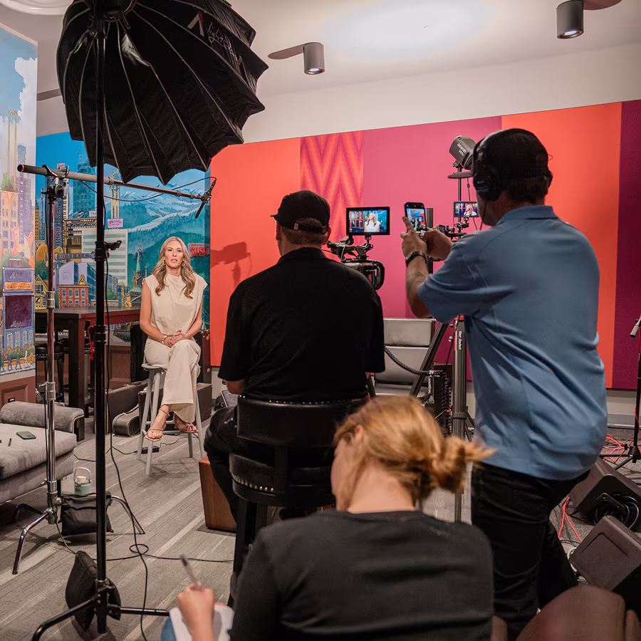 Woman in white outfit sitting on stool in studio, filmed by crew with cameras and lighting equipment.