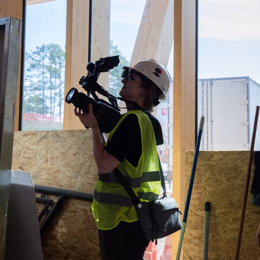 Person wearing hard hat and safety vest filming inside a construction site with a video camera.