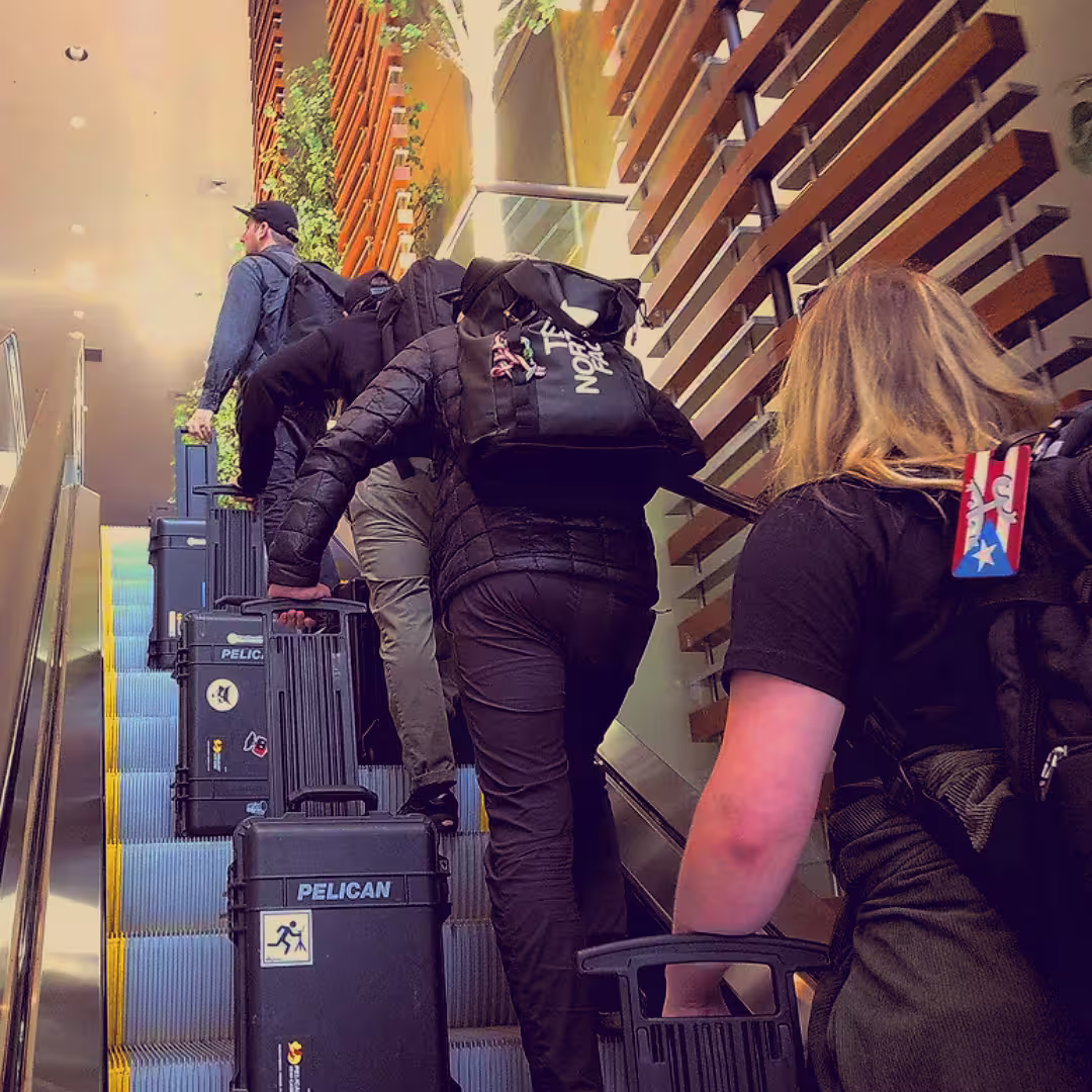 Four people carrying black Pelican cases up an escalator in a modern indoor space.