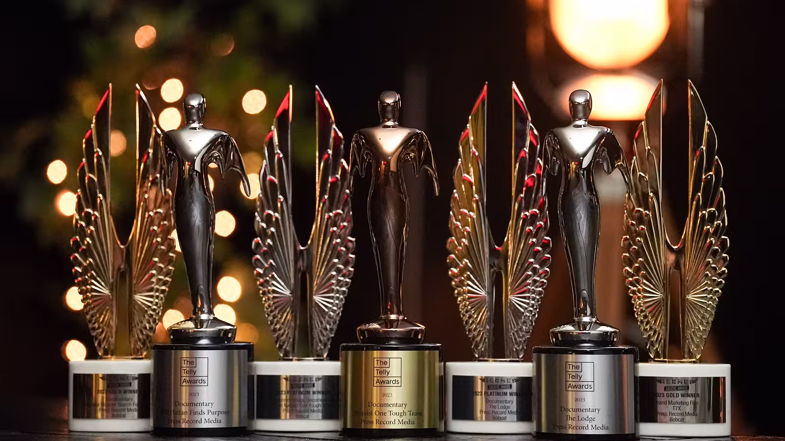 Several shiny Telly Awards trophies with winged figures on a dark table with blurred lights behind them.