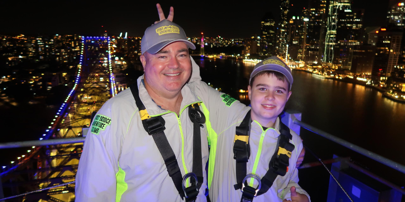 Father and son smiling on the Story Bridge at night, city lights in background.