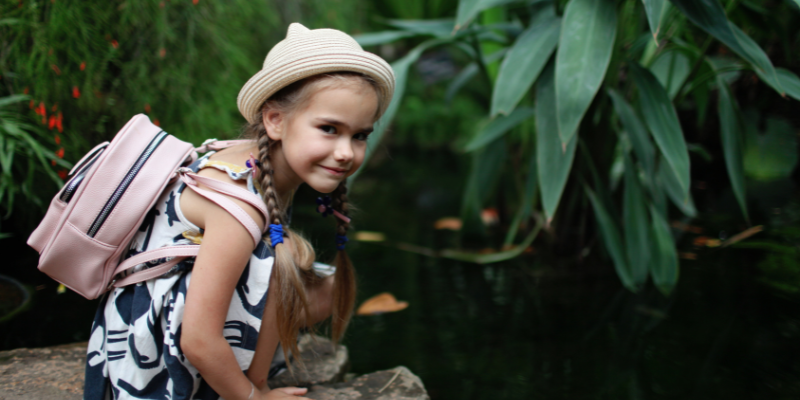 Young girl with hat and backpack smiling near pond.
