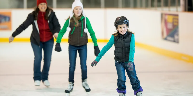 Family ice skating together indoors. A woman and two girls skate on the ice.