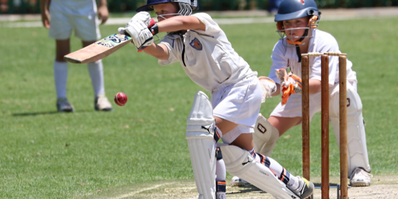 Young cricket player hitting a ball with another player behind the wickets.