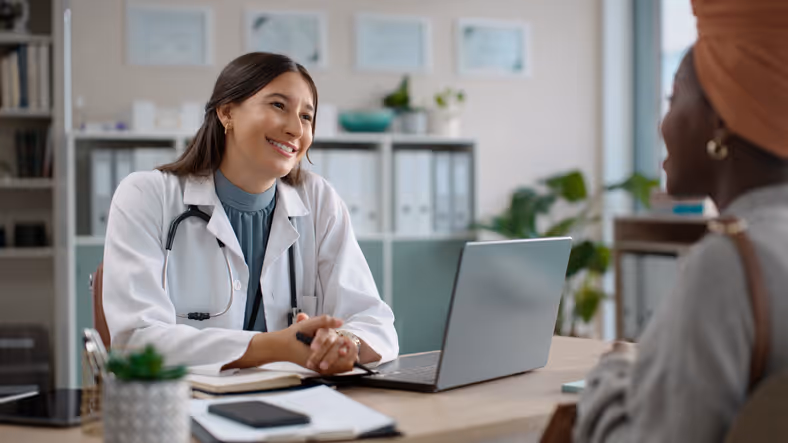Female doctor smiling and speaking with a patient across a desk in a medical office.