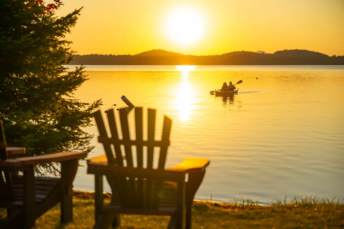Guests paddle a tandem kayak at sunset on Lake Clear, with an Adirondack Chair in the foreground.