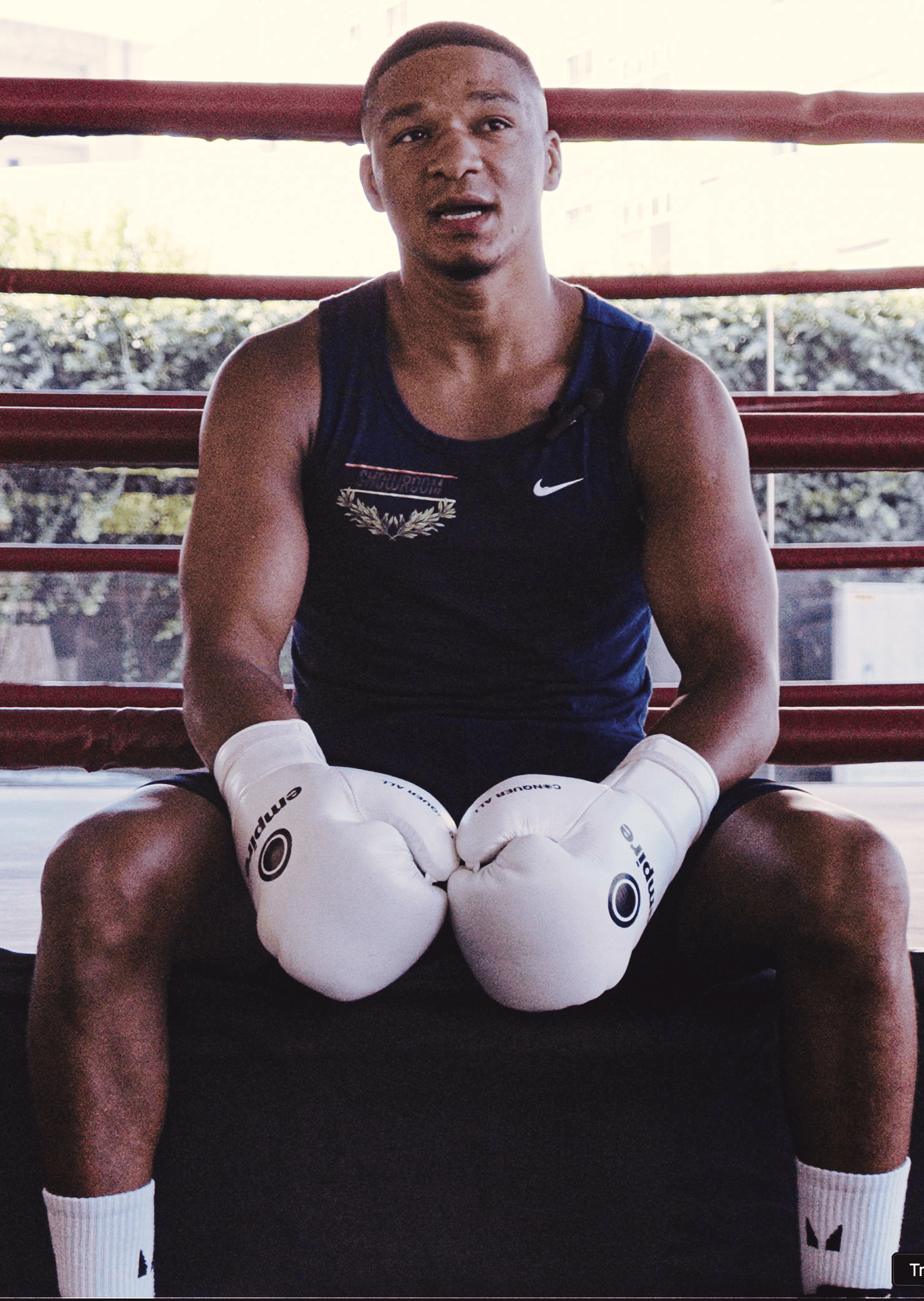 Boxer sitting in a ring wearing white gloves and a navy sleeveless shirt, resting with hands together.