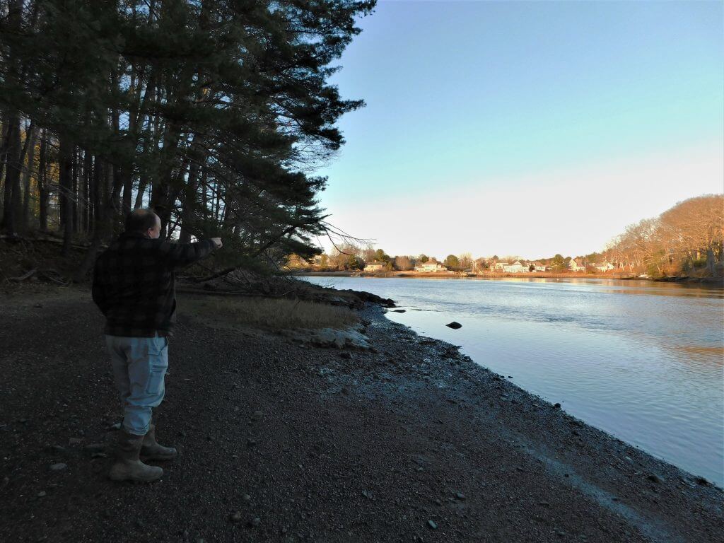 Person standing on rocky riverbank with pine trees and distant houses