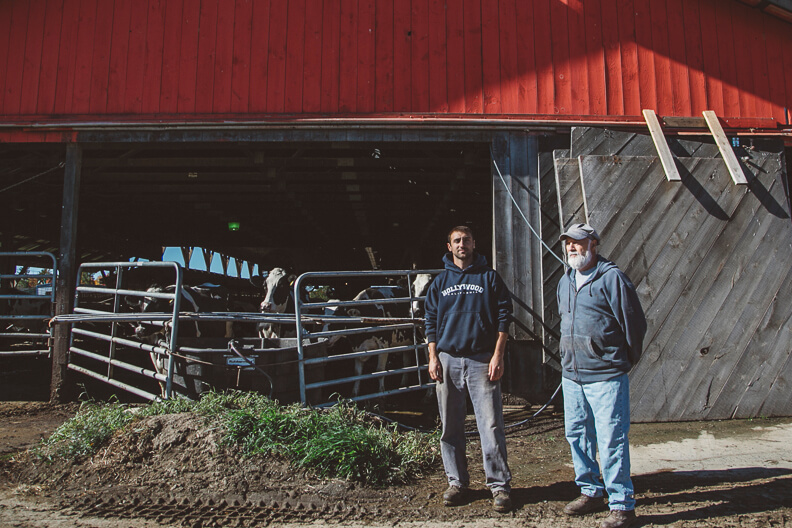 Two men standing in front of a red barn with livestock gates and cow